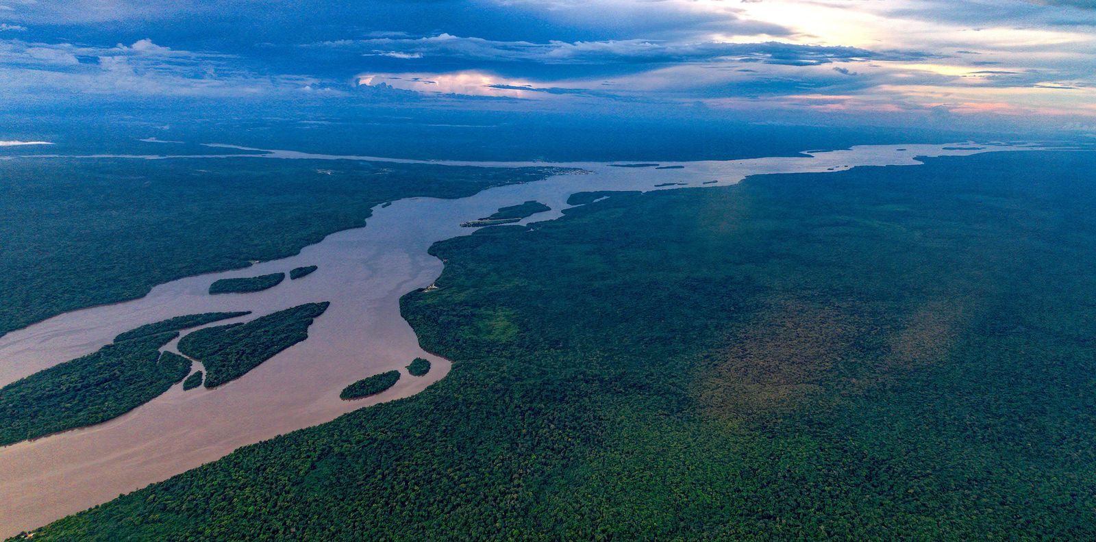 The Essequibo River at dusk from the air, winding wide and brown through unbroken forest near Bartica, with islands mid-river under a bank of cloud
