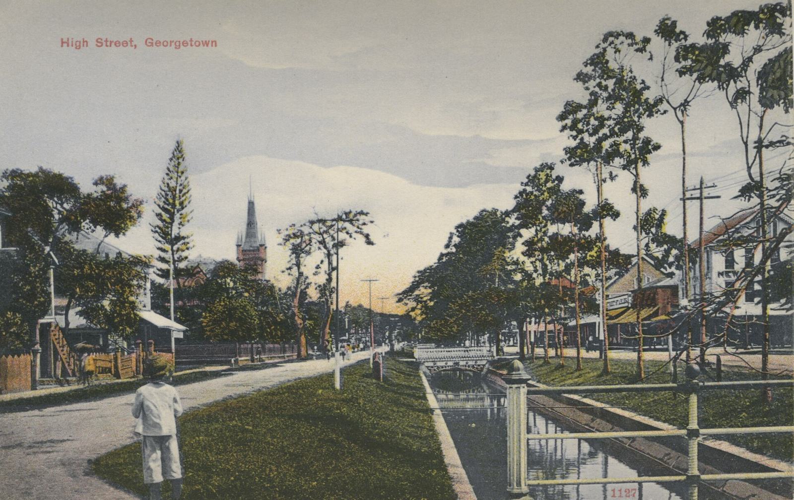 High Street, Georgetown, around 1910: the tree-lined avenue and its canal with white railings, a church spire beyond, a man in white standing at the roadside