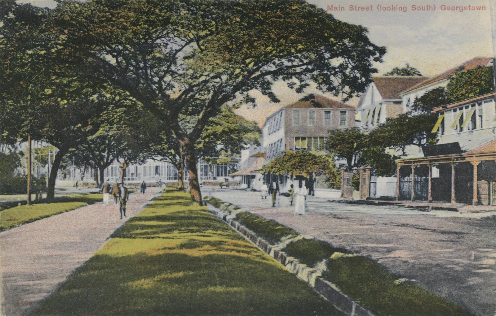 Main Street, Georgetown, looking south around 1910: a grass central walk under saman trees beside a canal, timber colonial houses and people strolling